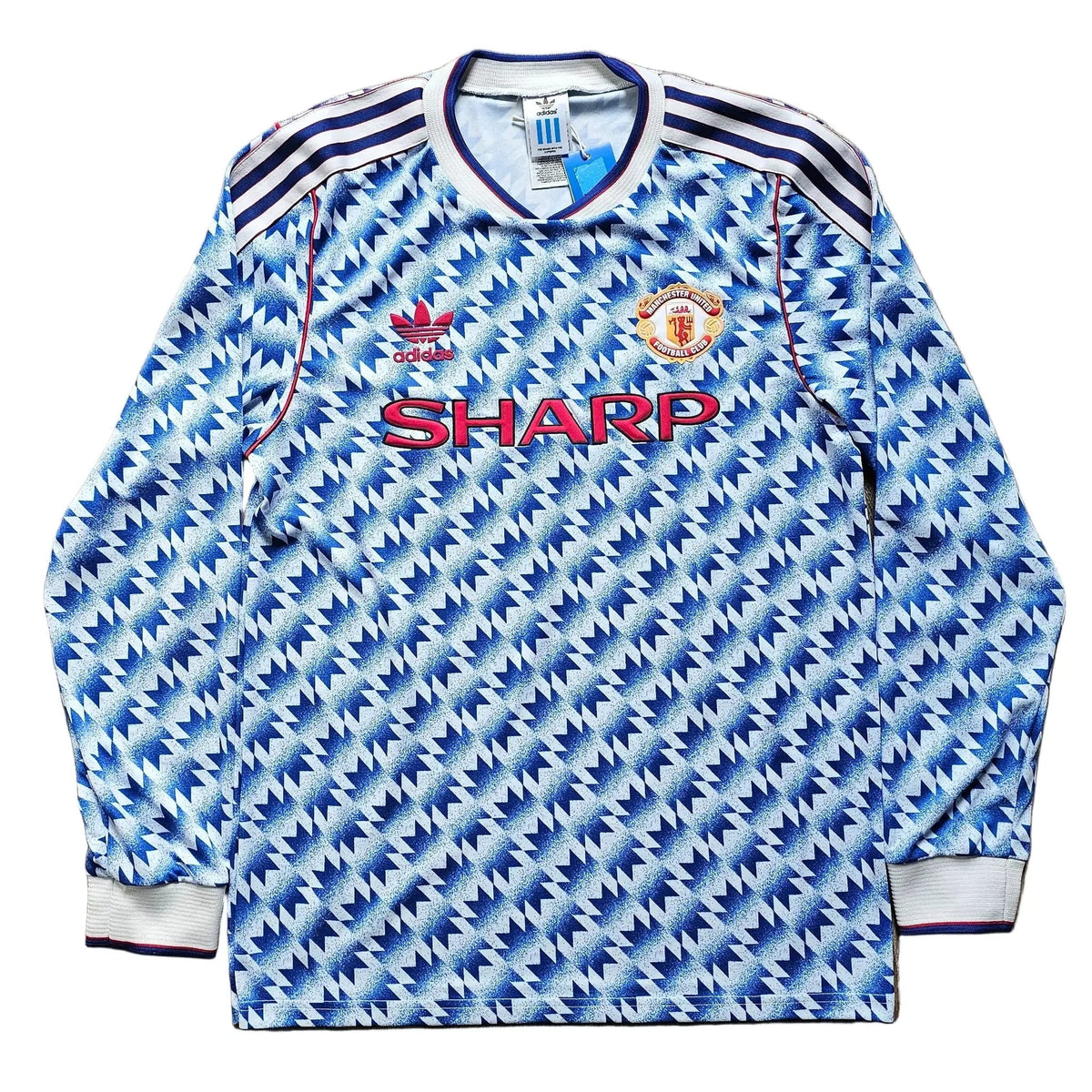 A long-sleeve Manchester United soccer jersey with a blue and white geometric pattern, laid flat on a light brown wooden floor. It features the red 'SHARP' sponsor logo across the chest, the Manchester United crest, and the Adidas logo.