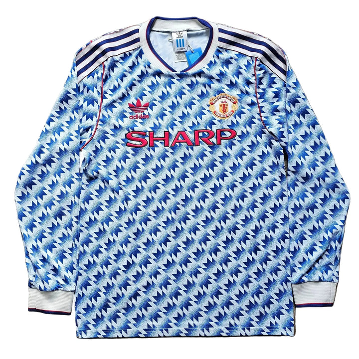 A long-sleeve Manchester United soccer jersey with a blue and white geometric pattern, laid flat on a light brown wooden floor. It features the red 'SHARP' sponsor logo across the chest, the Manchester United crest, and the Adidas logo.