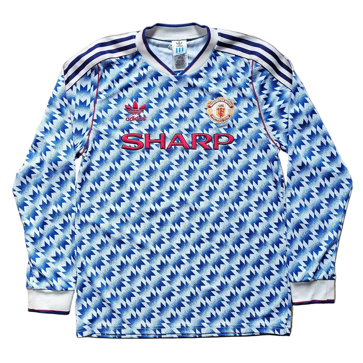 A vintage long-sleeve Manchester United soccer jersey by Adidas, featuring a blue and white patterned design with the red SHARP sponsor logo on the chest, laid flat on a wooden floor.
