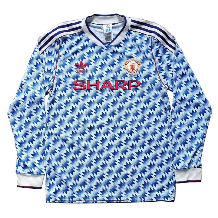 A vintage long-sleeve Manchester United soccer jersey by Adidas, featuring a blue and white patterned design with the red SHARP sponsor logo on the chest, laid flat on a wooden floor.