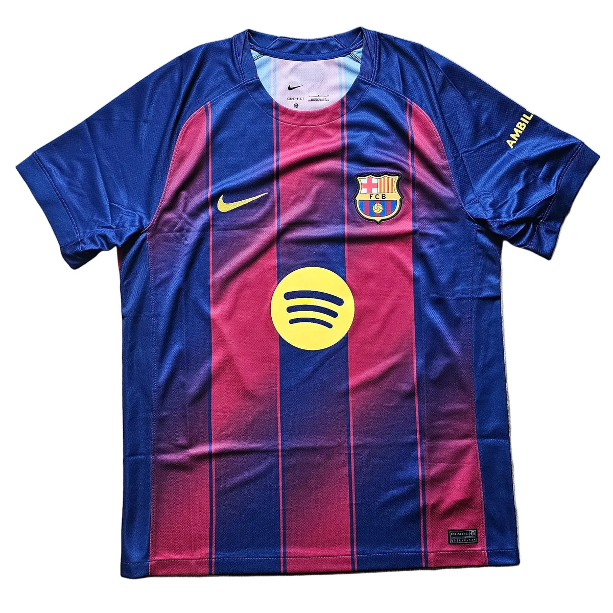 A Nike FC Barcelona home soccer jersey with blue and magenta vertical stripes, featuring the yellow Spotify sponsor logo on the front, laid flat on a light wood floor.