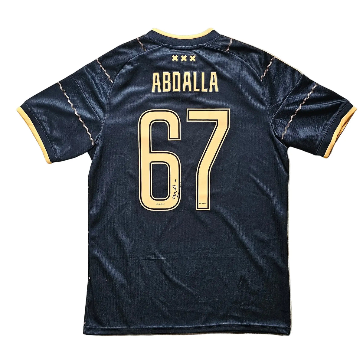 A black soccer jersey with 'ABDALLA' and the number '67' in gold on the back, laid flat on a light wood floor.