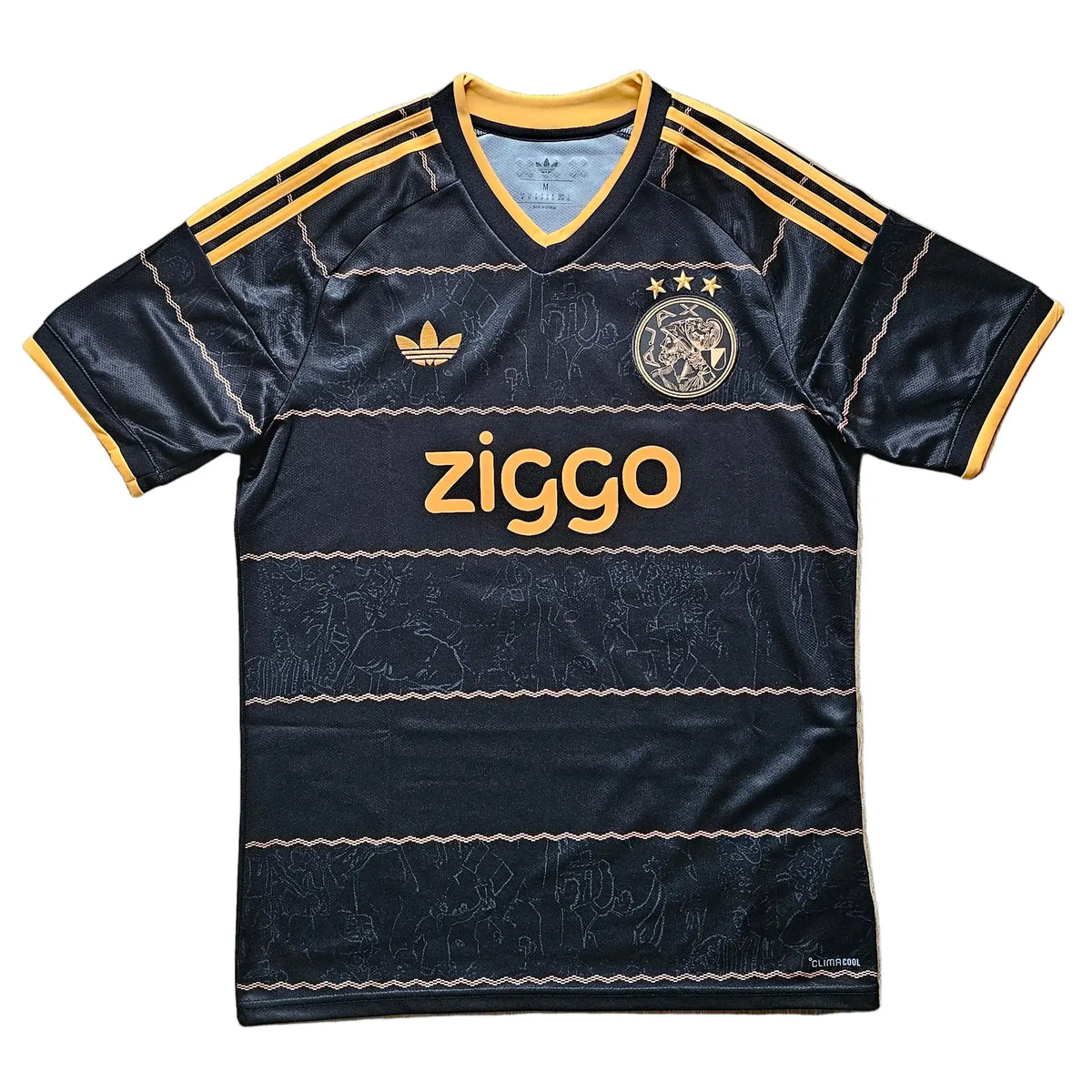 A black Ajax Amsterdam soccer jersey by Adidas, featuring yellow stripes on the shoulders, a yellow ziggo sponsor logo on the chest, and a yellow Ajax club crest, laid flat on a light wood plank background.