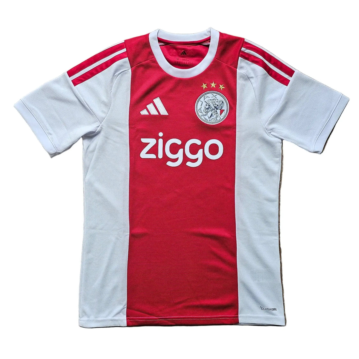 A red and white short-sleeve soccer jersey laid flat on a wooden floor. The jersey features a vertical red stripe down the center, white side panels and sleeves, red shoulder stripes, the Adidas logo on the right chest, a circular club crest on the left chest, and 'ziggo' sponsor text across the front.