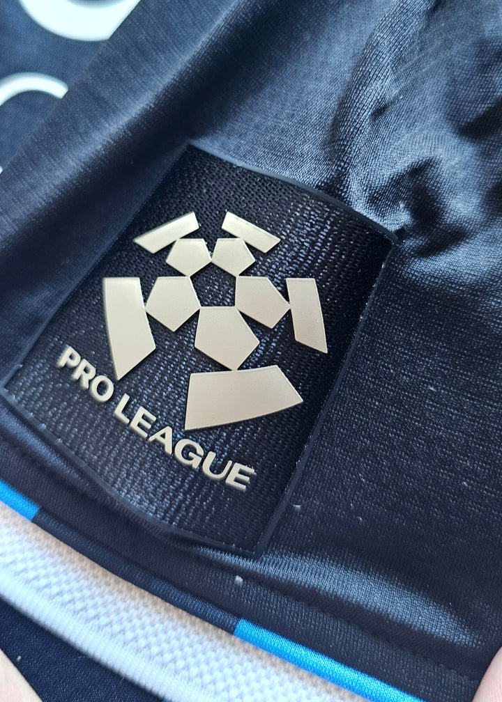 Pro League sleeve badge in gold on black, above the white and blue cuff of the Club Brugge 2026-27 home shirt, Koren 85