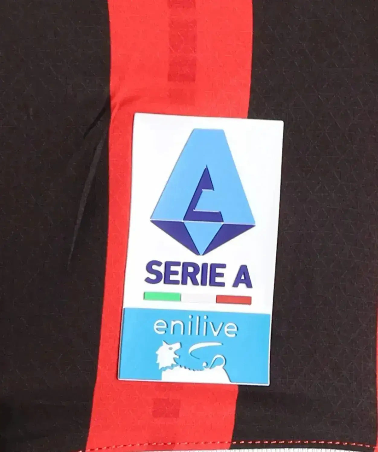 Crest detail showing Serie A logo and enilive brand on red and black football shirt.