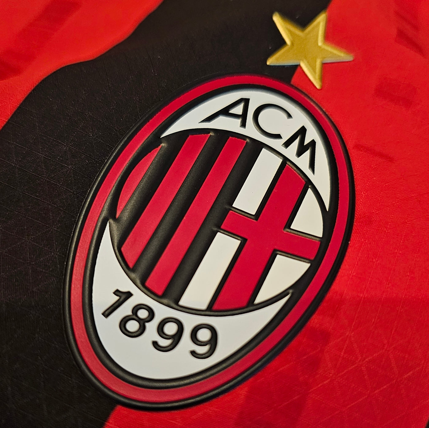 AC Milan crest on a red and black football shirt, oval badge with black and red stripes and a red cross, ACM 1899 text and a single gold star above.