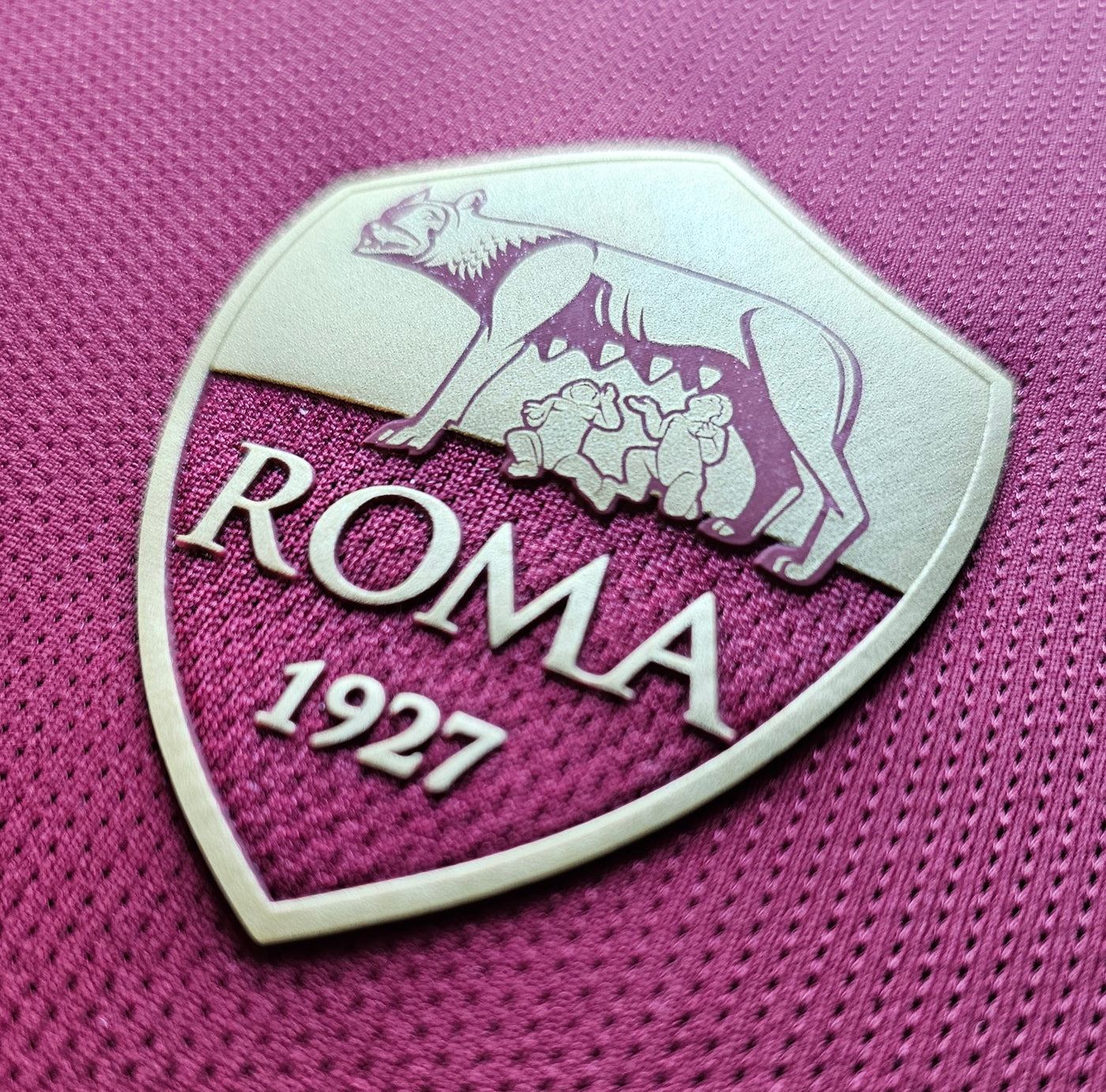 AS Roma crest close up on a magenta football shirt with a metallic beige shield showing the she wolf with Romulus and Remus and the text ROMA 1927 on textured mesh.
