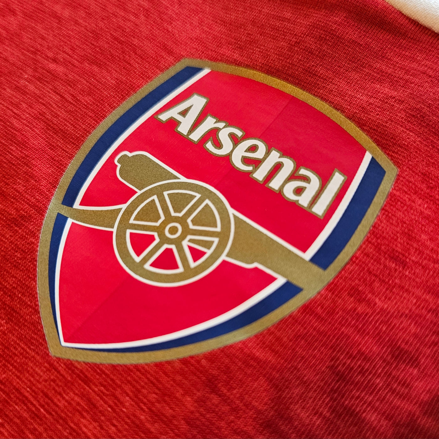 Arsenal crest on a deep red football shirt, red shield with gold cannon and gold edged border.