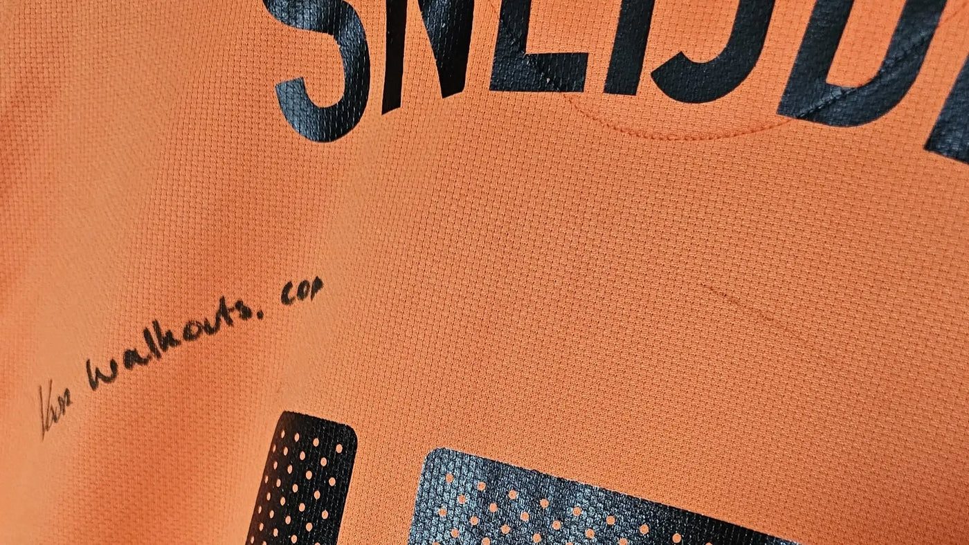 Close-up of an orange football shirt with black text "VOOR WALKOUTS.COM" and patterns.