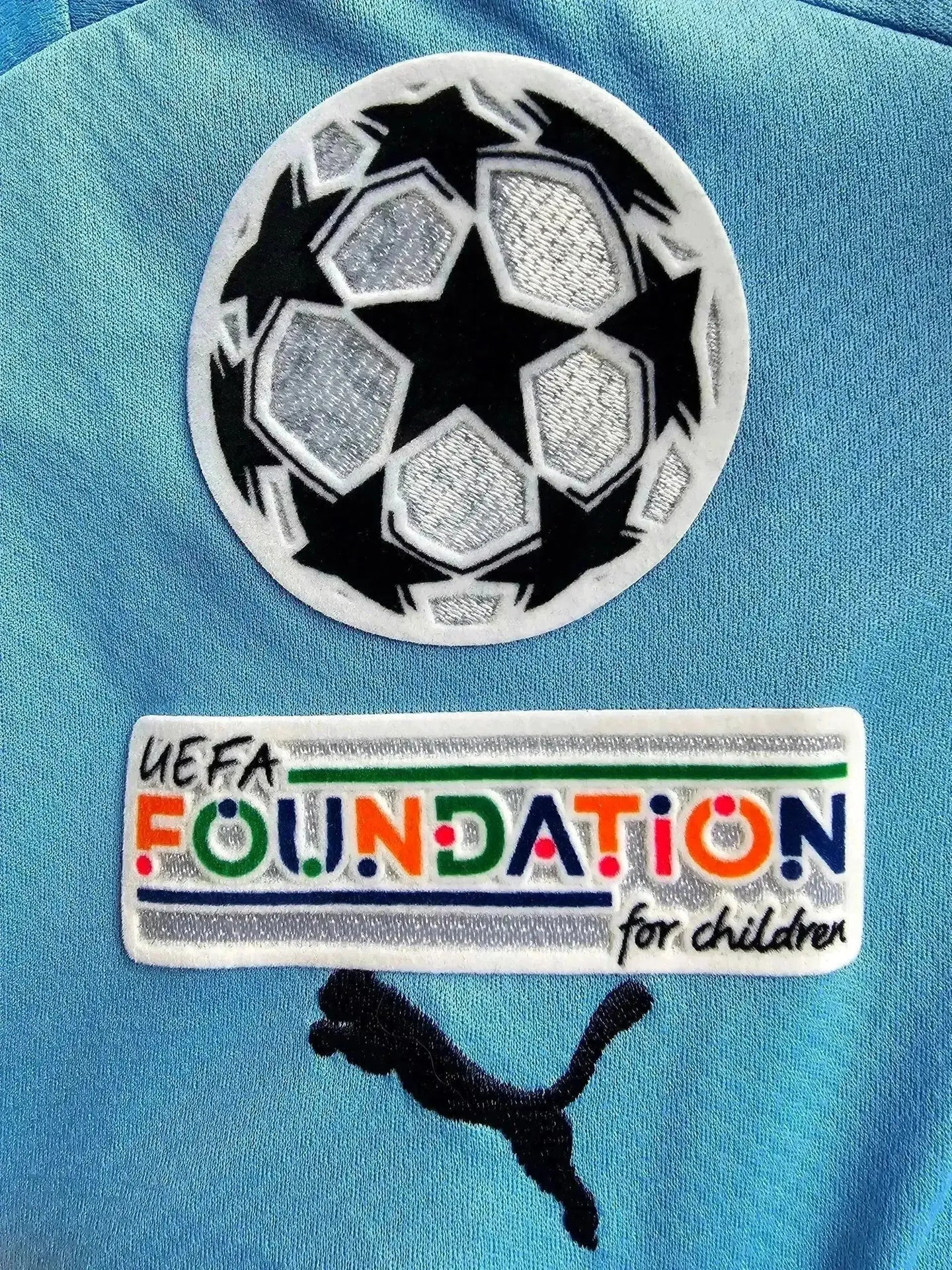 Crest detail showing UEFA Champions League ball and UEFA FOUNDATION for children patch on blue fabric.