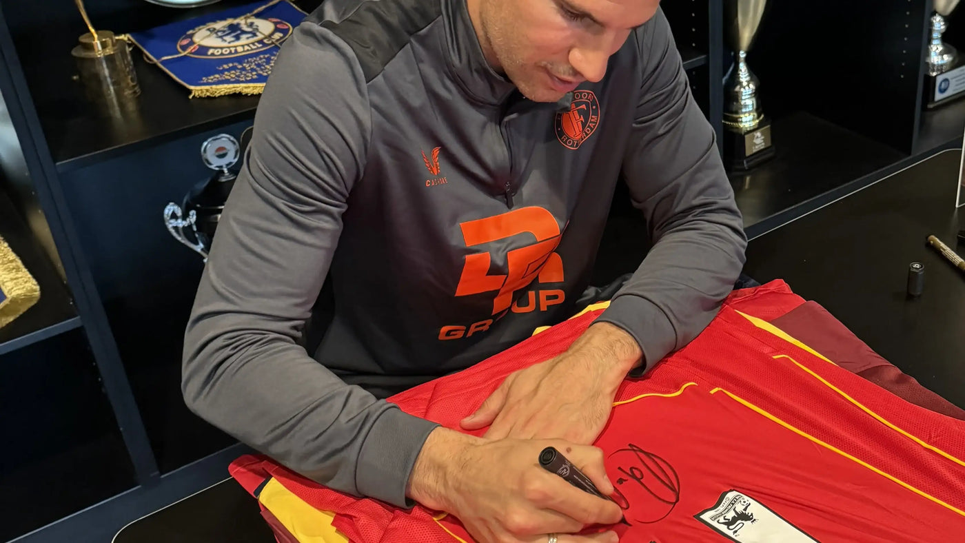 Robin van Persie signing a red Arsenal football shirt with trophies and awards in the background.