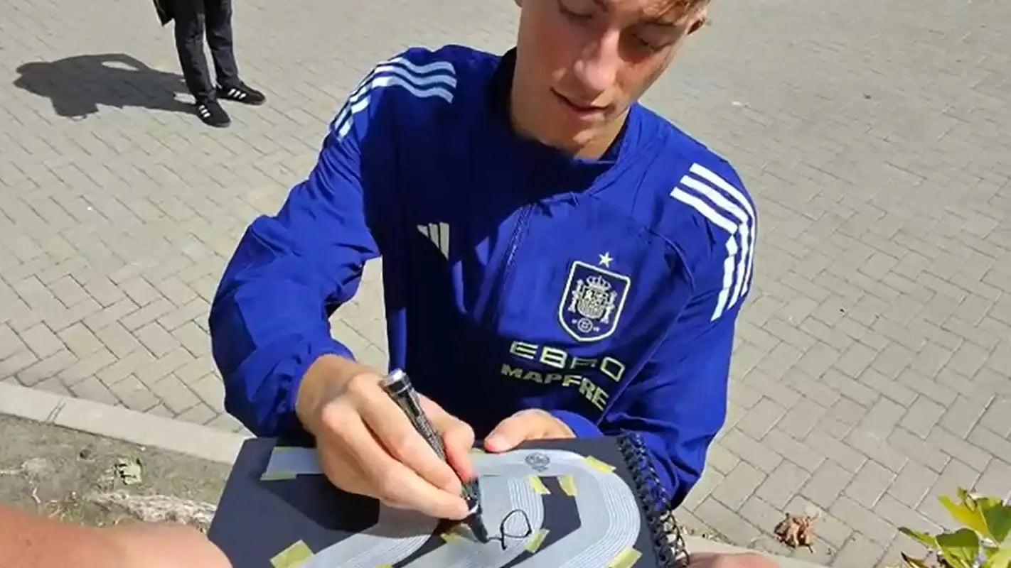 Dean Huijsen in a blue sports jersey signing an item outdoors on a paved surface.