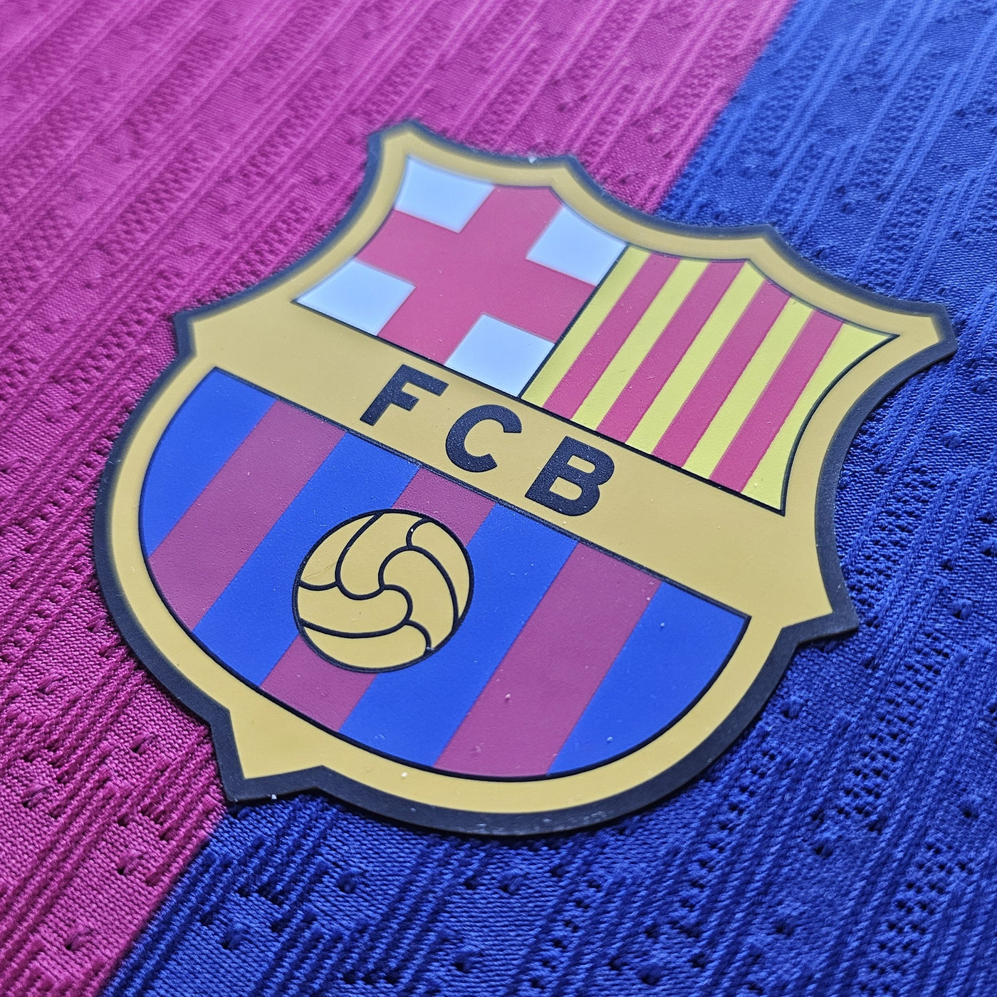 FC Barcelona crest close up on a magenta and deep blue football shirt with a textured knit, yellow edged badge showing the red cross, red and yellow stripes and FCB lettering.