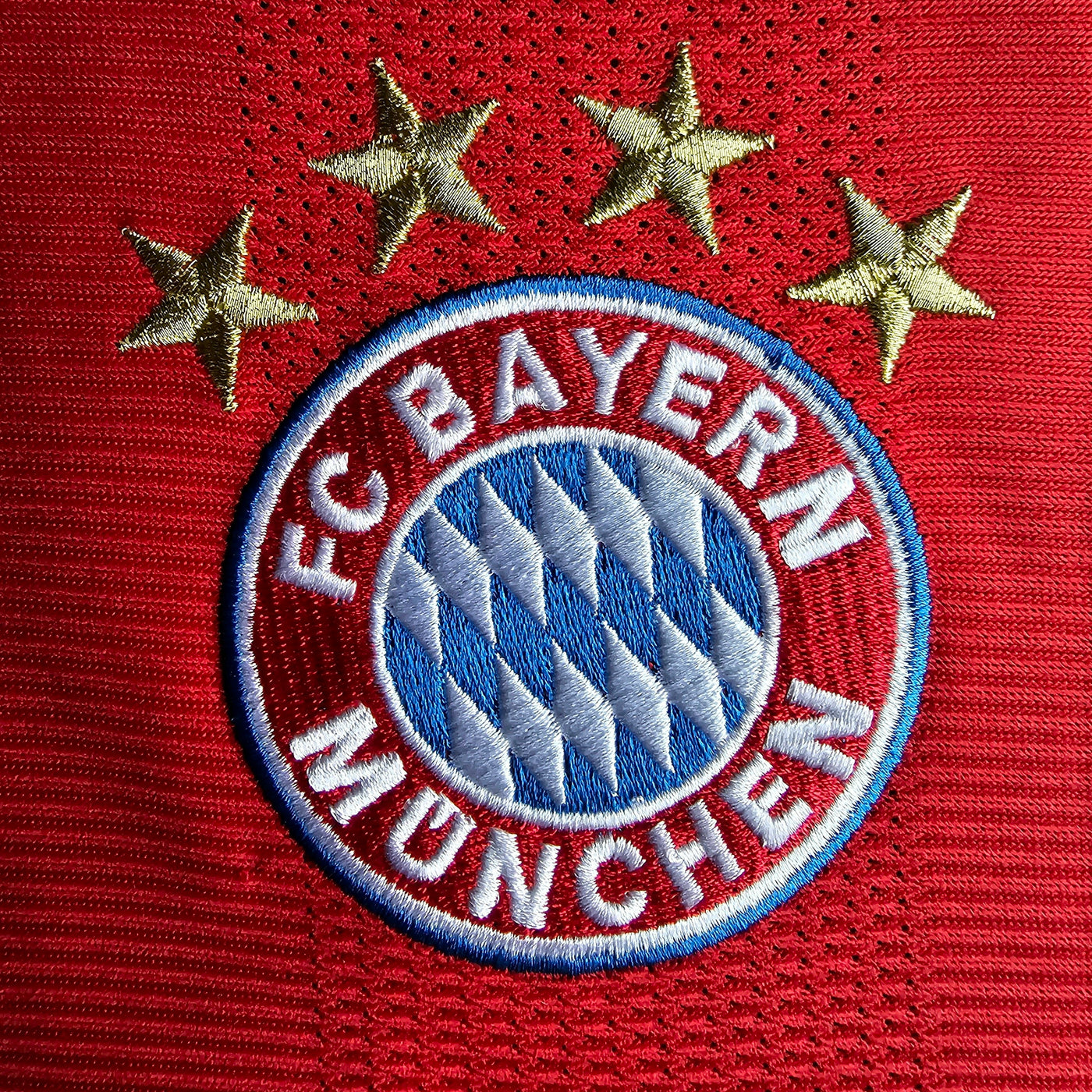 FC Bayern München crest close up on a red football shirt, embroidered roundel with blue and white diamonds and four gold stars above.