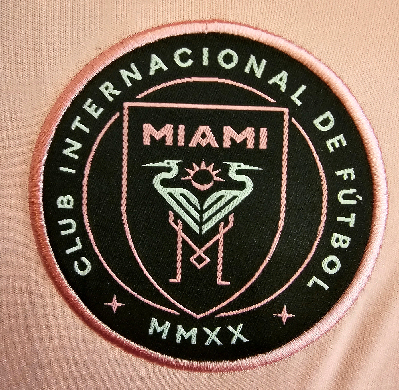 Inter Miami CF crest on a pale pink football shirt, black roundel with a pink border, stylized herons and sun, Club Internacional de Fútbol and MMXX text.