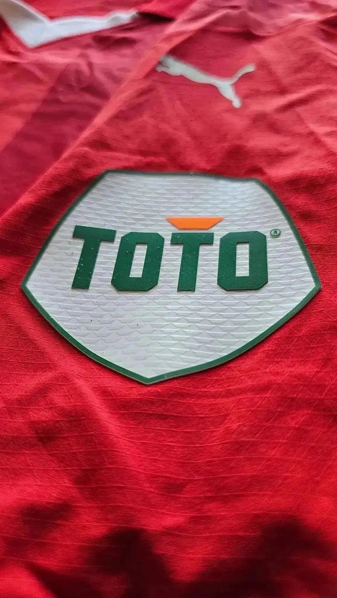 Close-up of the TOTO sponsor logo on a red soccer jersey, featuring the trademark design.