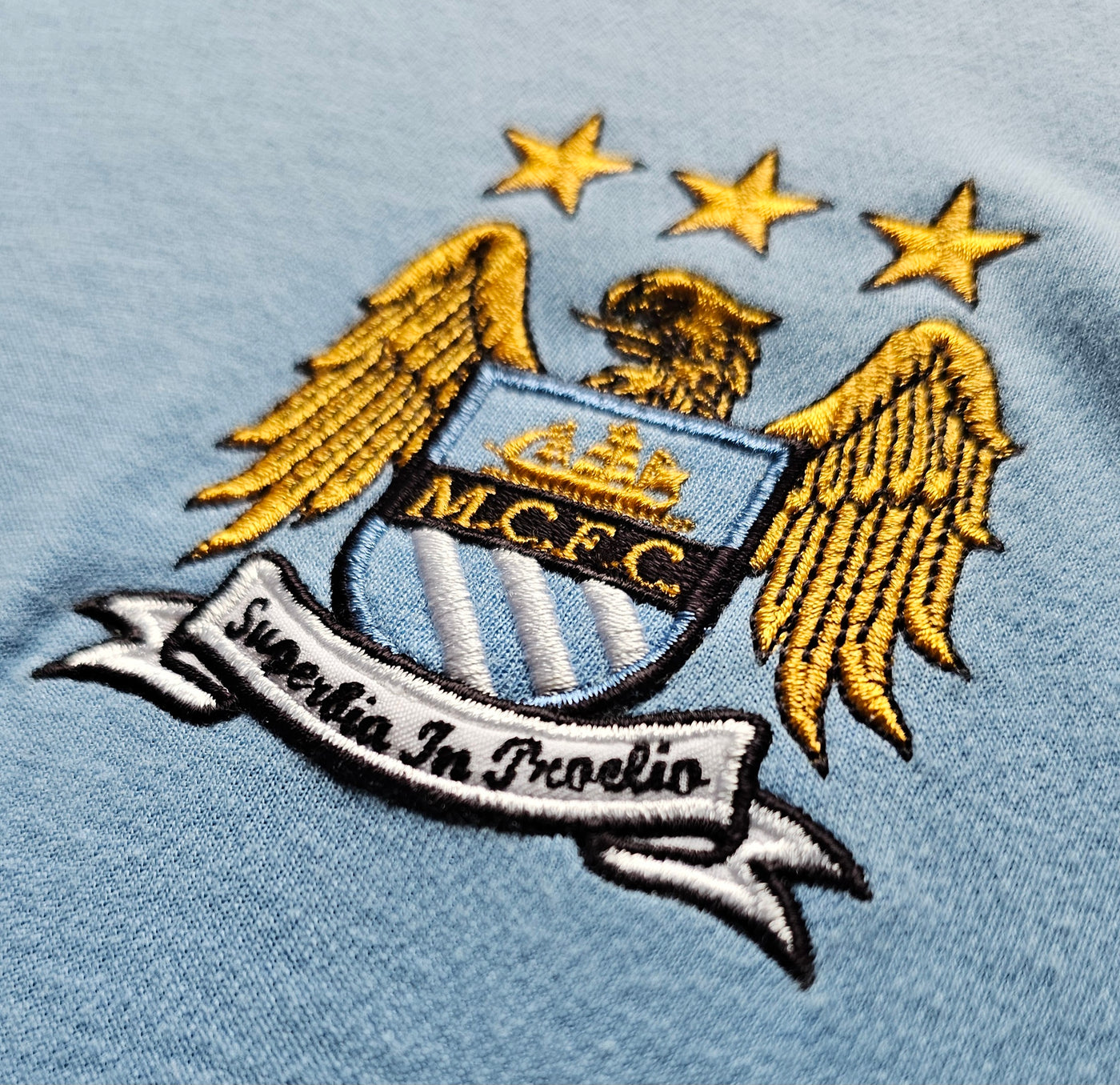 Manchester City crest on a light blue football shirt, embroidered shield with golden eagle wings, three gold stars, MCFC initials and the Superbia In Proelio banner.