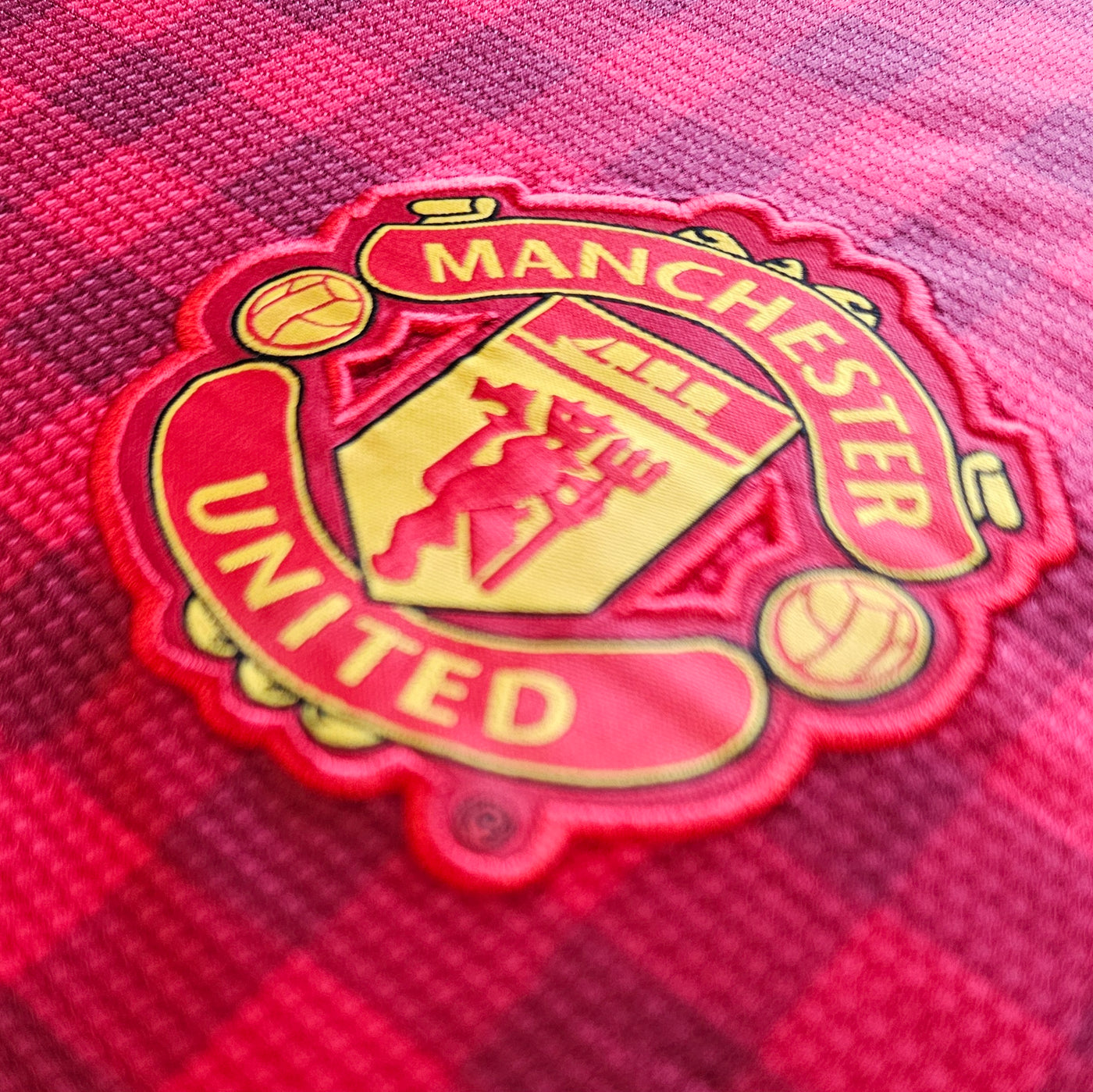 Manchester United crest on a red football shirt with a subtle check pattern, red and yellow badge with black outline and stitched detail.