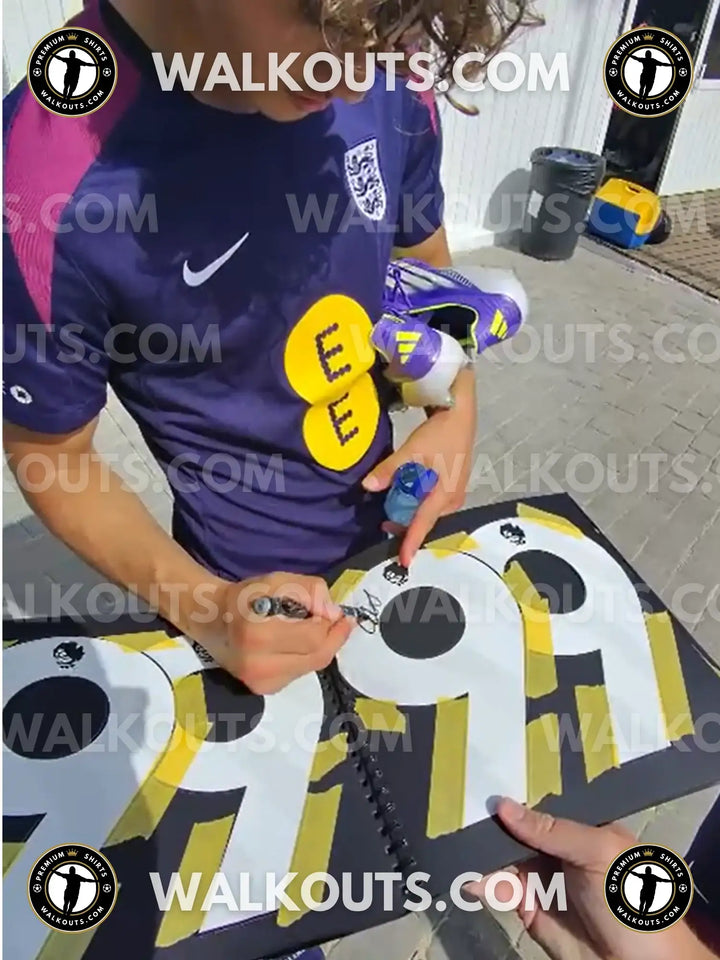 Max Dowman signing a black folder with yellow and black shirt numbers 99 visible.