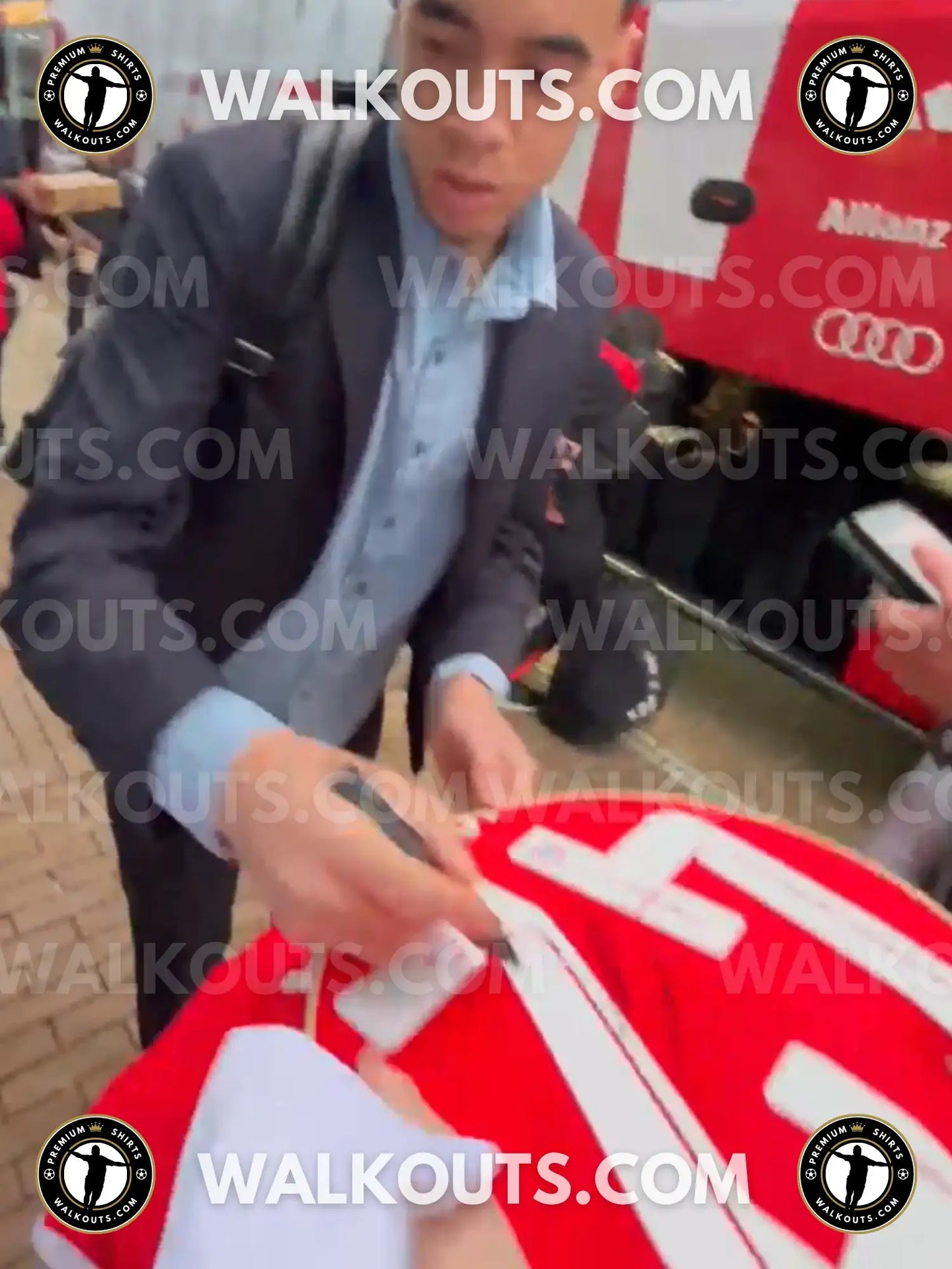 Jamal Musiala signing a red and white Bayern Munich jersey with 'WALKOUTS.COM' branding.