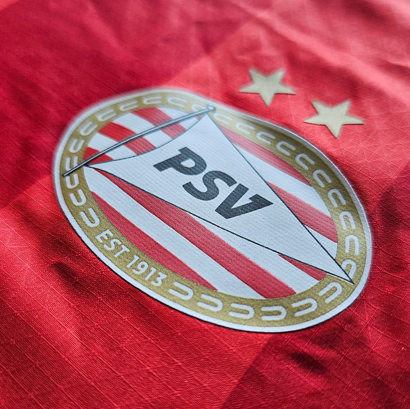 PSV Eindhoven crest on a red football shirt, oval badge with red and white stripes, gold chain border and two gold stars above.