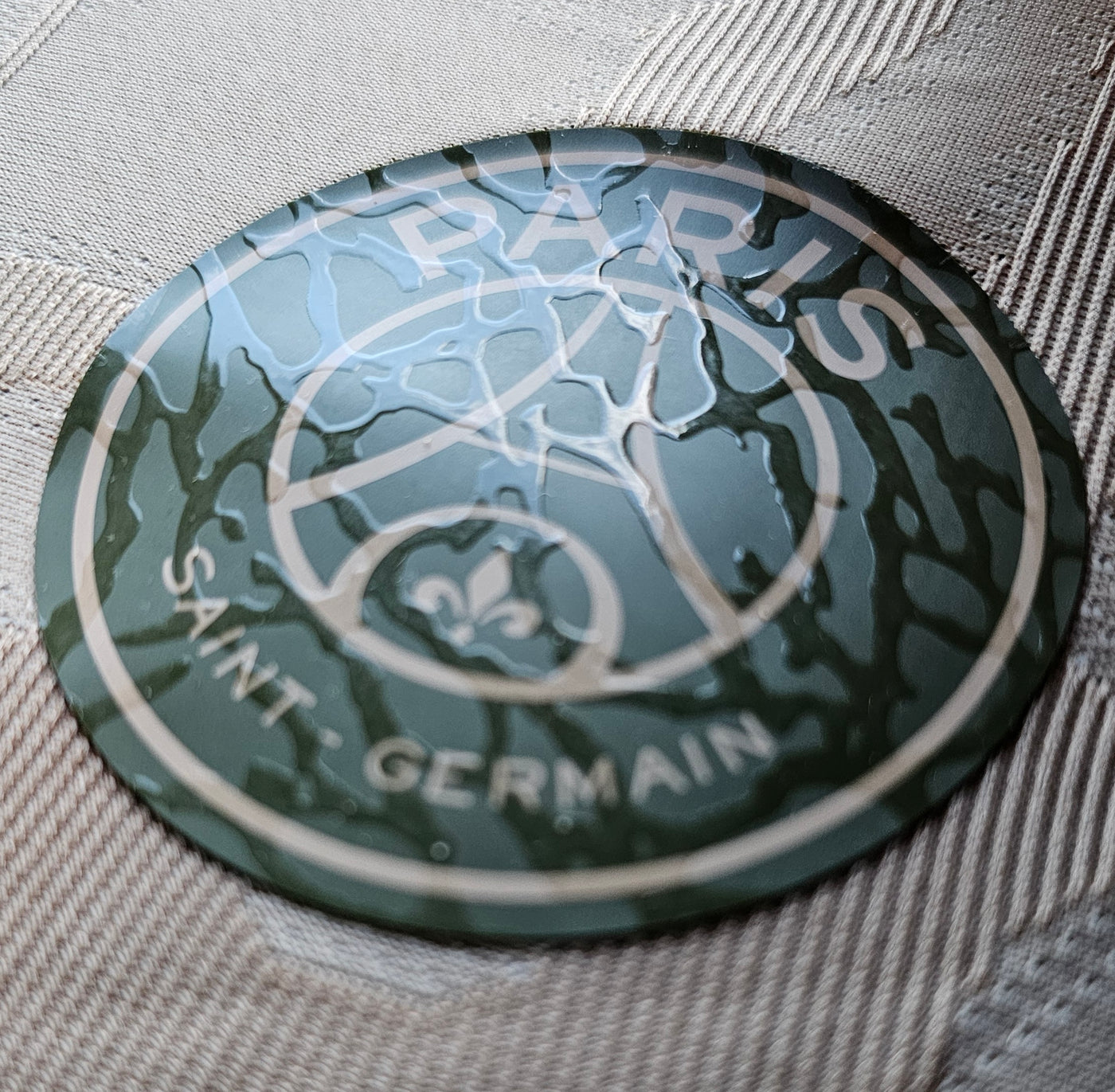 Paris Saint Germain tonal crest on a beige fabric patch, dark green roundel with glossy raised details of the Eiffel Tower and fleur de lis.