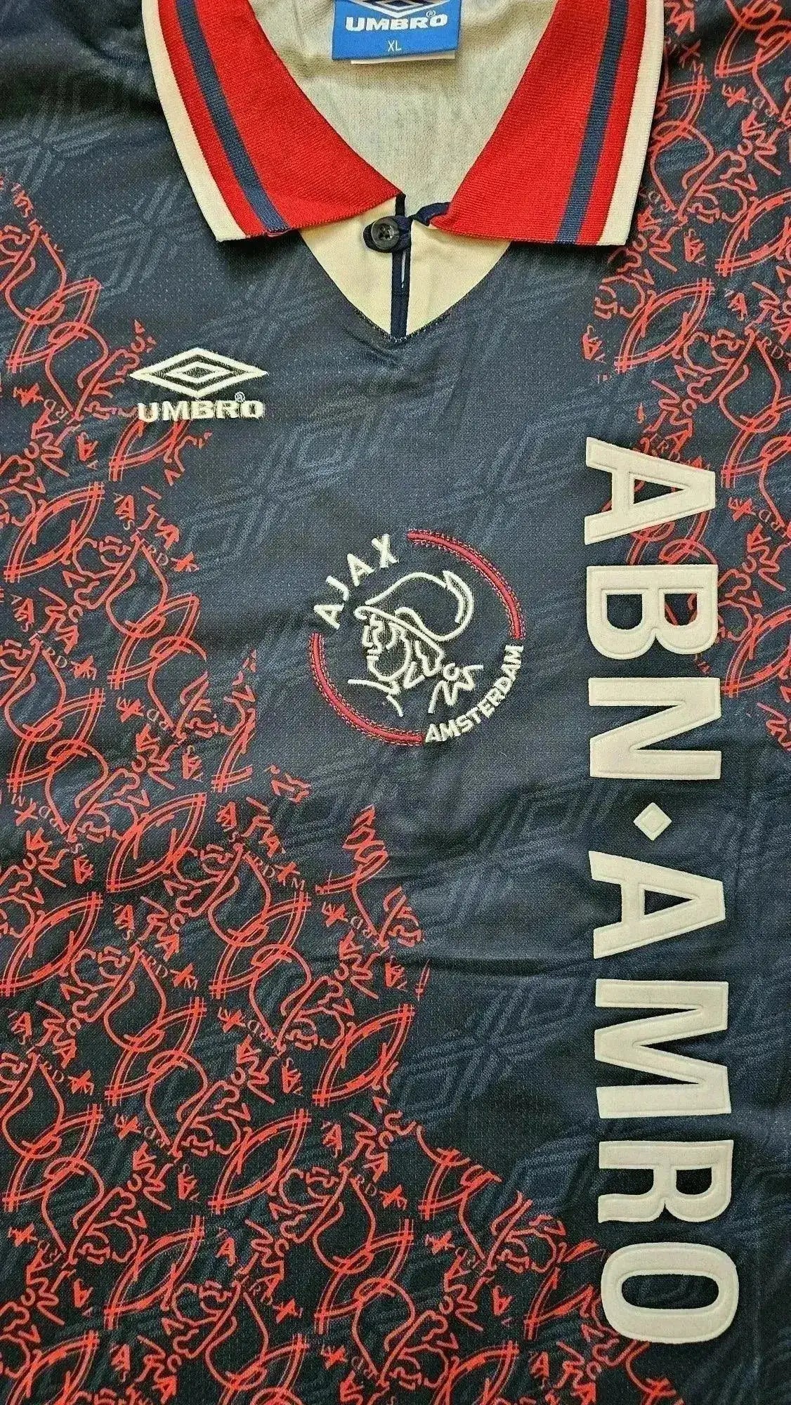 Front view showing Ajax crest, ABN AMRO sponsor, and intricate red pattern on a dark base.
