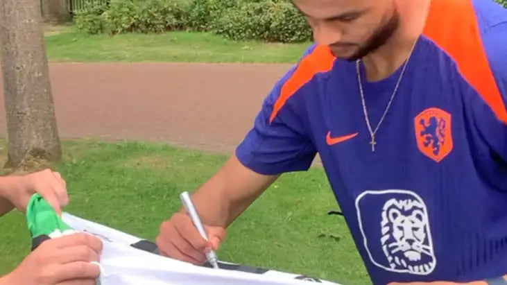 Cody Gakpo in a blue sports jersey with orange accents signing a white item outdoors.