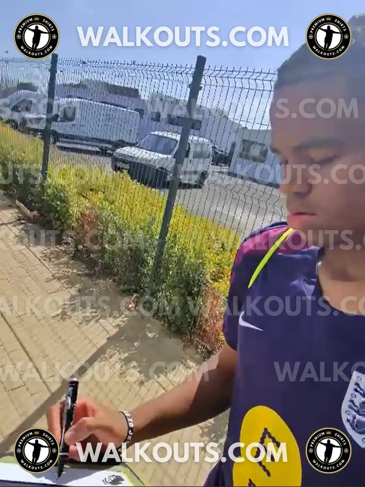 Rio Ngumoha signing a Liverpool F.C. football shirt with a marker on a sunny day.
