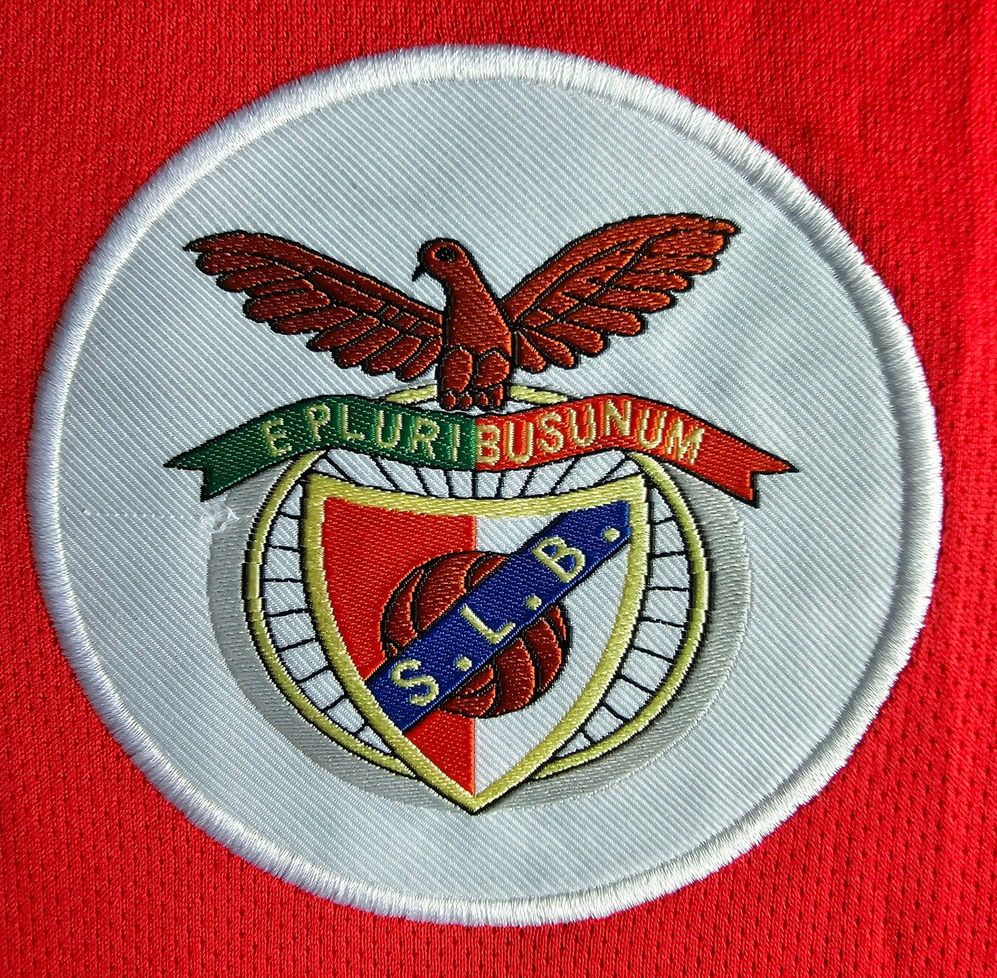 S.L. Benfica crest close up on a bright red football shirt with a white oval patch, an eagle above a green and red E Pluribus Unum banner, and a shield with red panel, golden wheel and blue SLB diagonal.