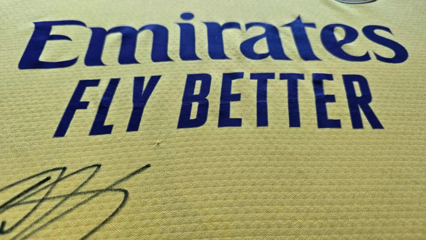 Close-up of 'Emirates FLY BETTER' with signature detail on yellow football shirt.