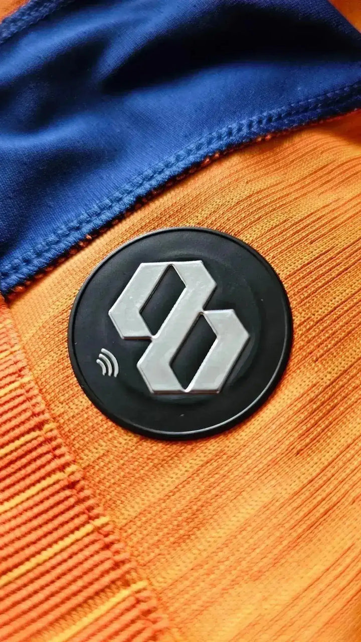 Close-up of black and silver logo patch on an orange football shirt with blue trim.