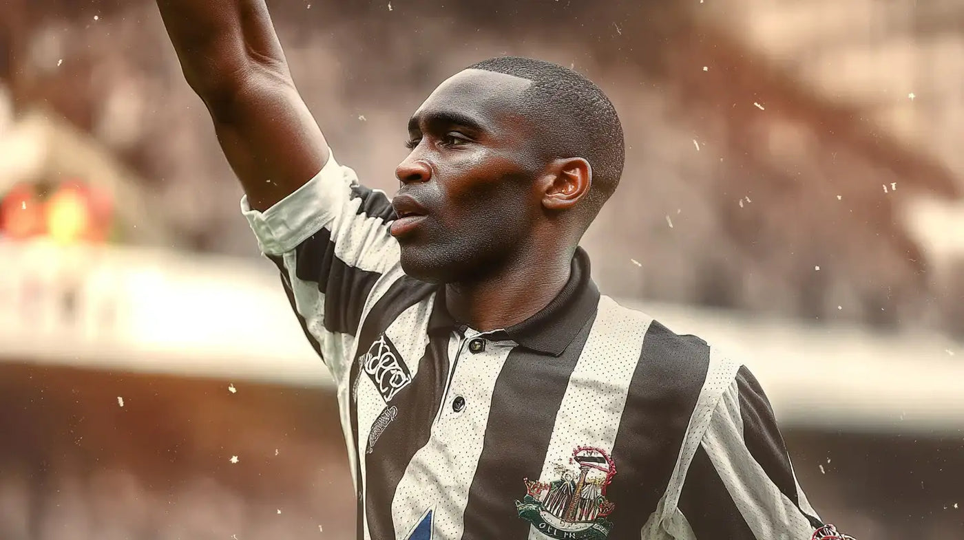 Andy Cole raising his arm in a Newcastle United black and white striped football shirt with a black collar, with a blurred crowd in the background.