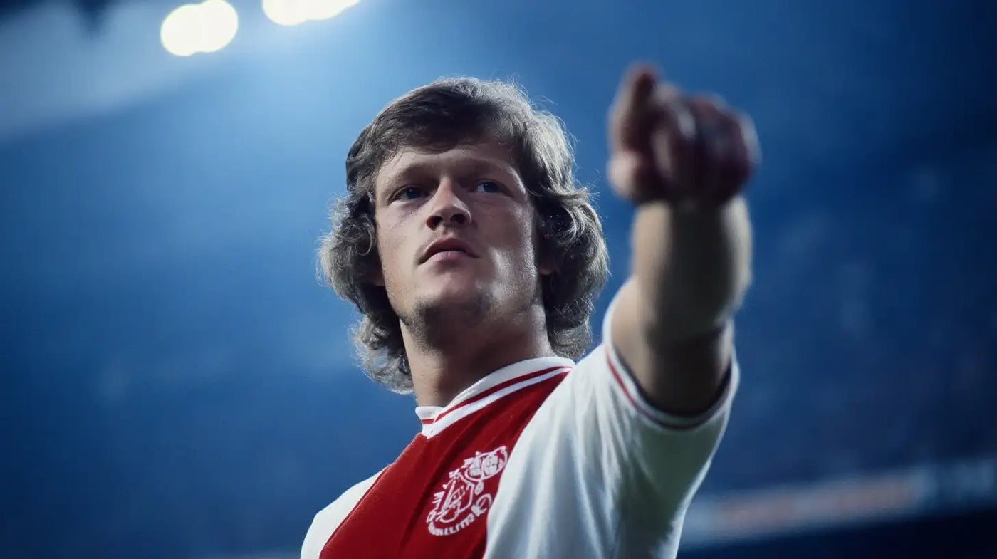 Arie Haan points ahead in the classic Ajax red and white kit, vintage stadium scene under bright lights.