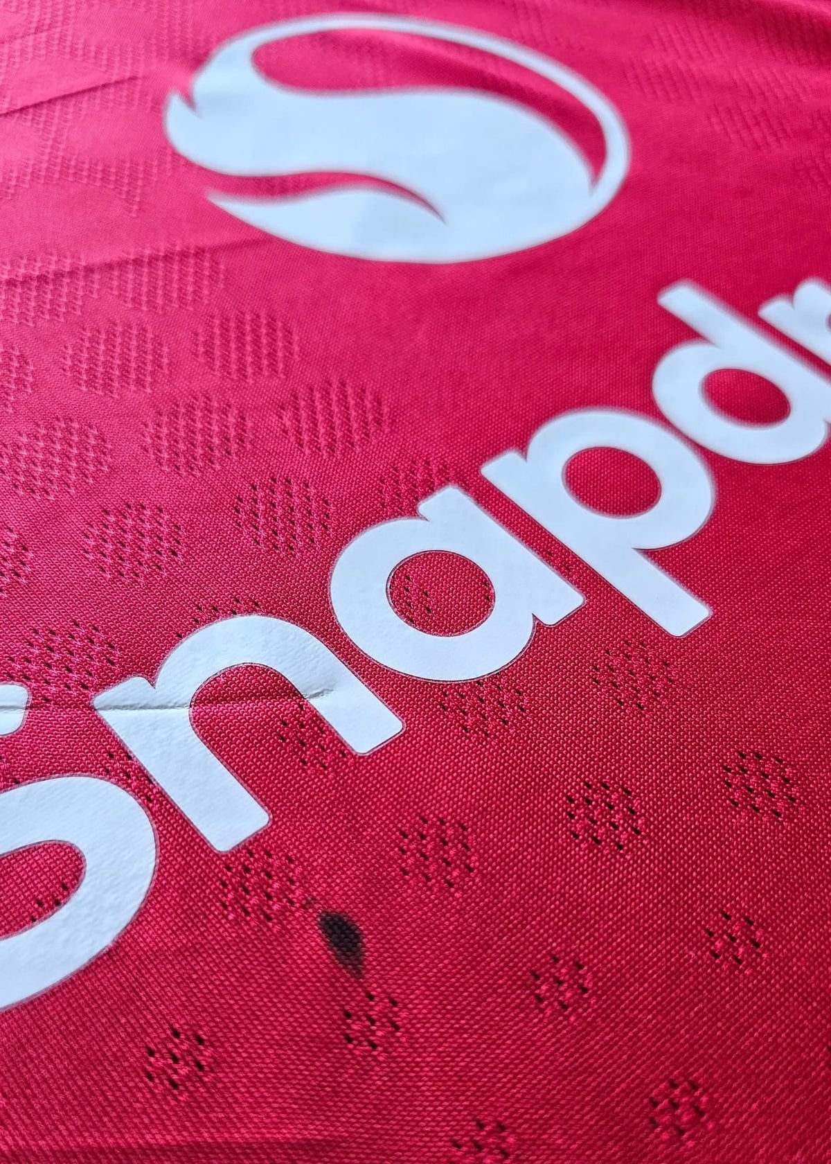 Close-up view of red football shirt featuring white Snapdr logo and textural details.