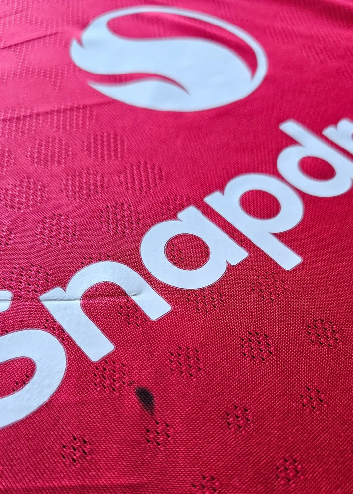 Close-up view of red football shirt featuring white Snapdr logo and textural details.