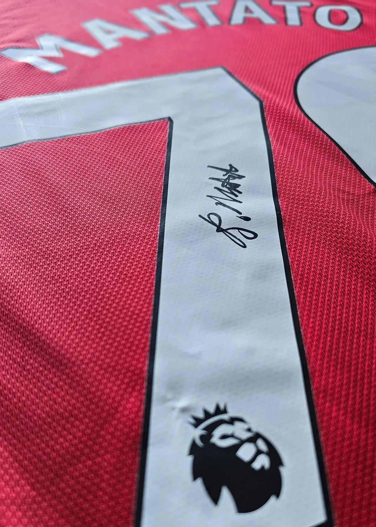 Close-up of the white No. 70 with a black-ink autograph and the Premier League crest patch beneath it, on Bendito Mantato's red Manchester United shirt.
