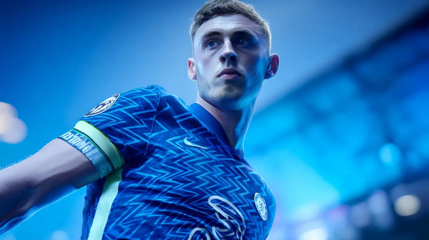 Cole Palmer looks over his shoulder under cool blue arena lighting, wearing Chelsea’s patterned blue football shirt with the captain’s armband and the club crest visible.