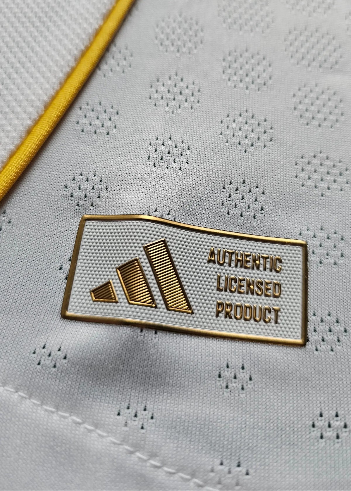 Close-up of the adidas Authentic Licensed Product tag near the hem, beside the yellow trim of the Real Madrid shirt.