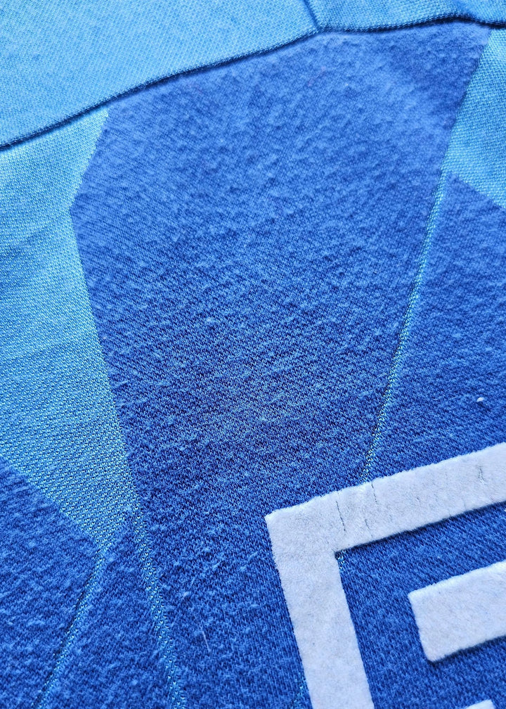 Close-up of blue textured fabric with a geometric pattern and a white embroidered letter E on the lower right.