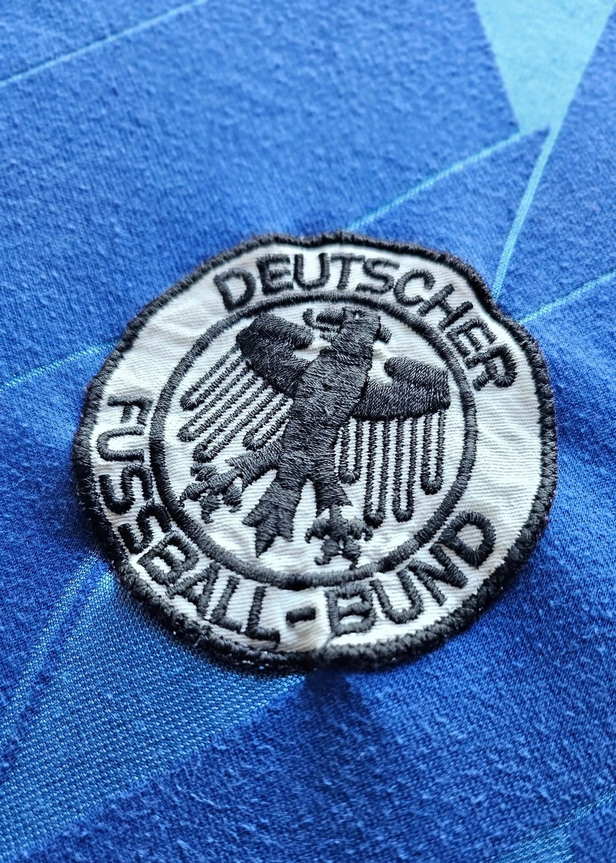 Round DFB (Deutscher Fussball-Bund) federation patch with an embroidered eagle emblem, stitched onto Eike Immel's blue West Germany 1987-88 shirt.