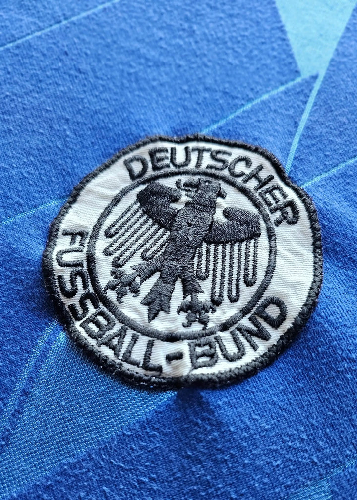 Round DFB (Deutscher Fussball-Bund) federation patch with an embroidered eagle emblem, stitched onto Eike Immel's blue West Germany 1987-88 shirt.