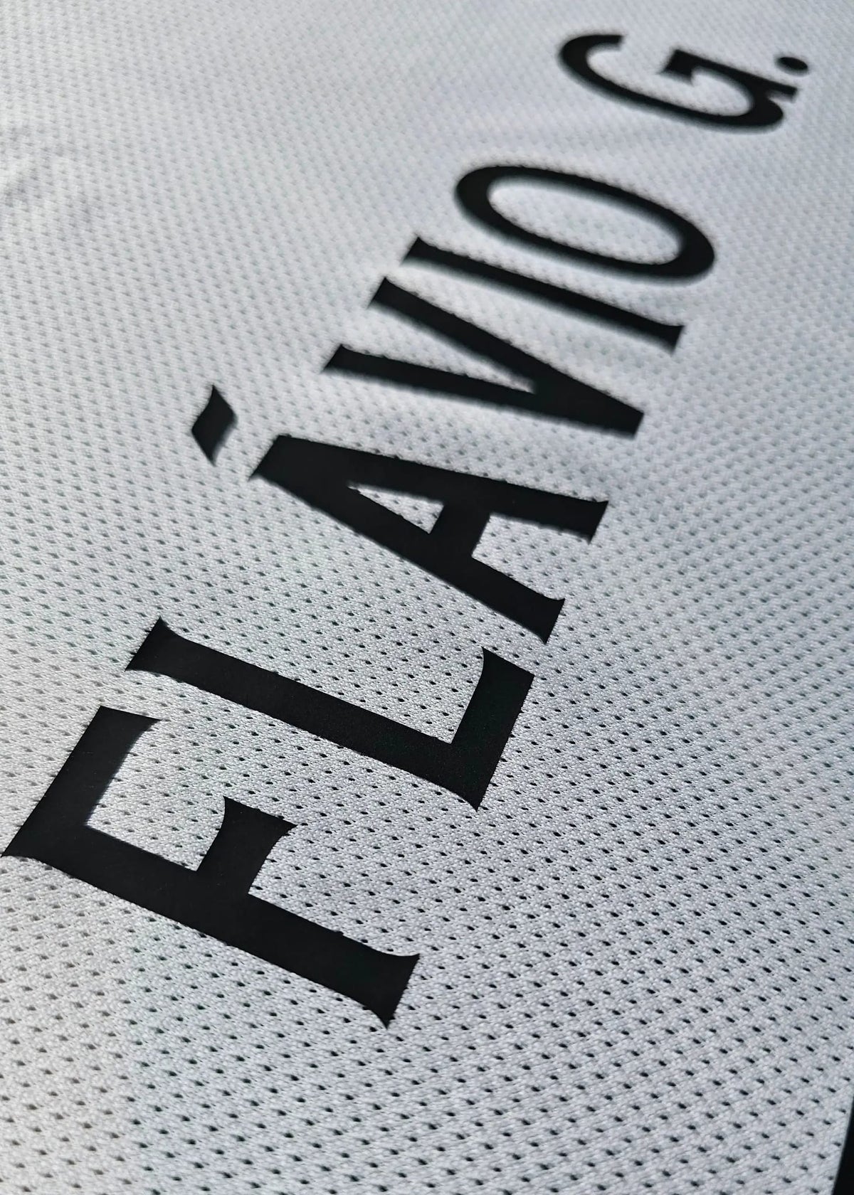 Close-up of a white perforated athletic jersey with bold black text reading 'FLÁVIO G.' printed diagonally across the fabric.