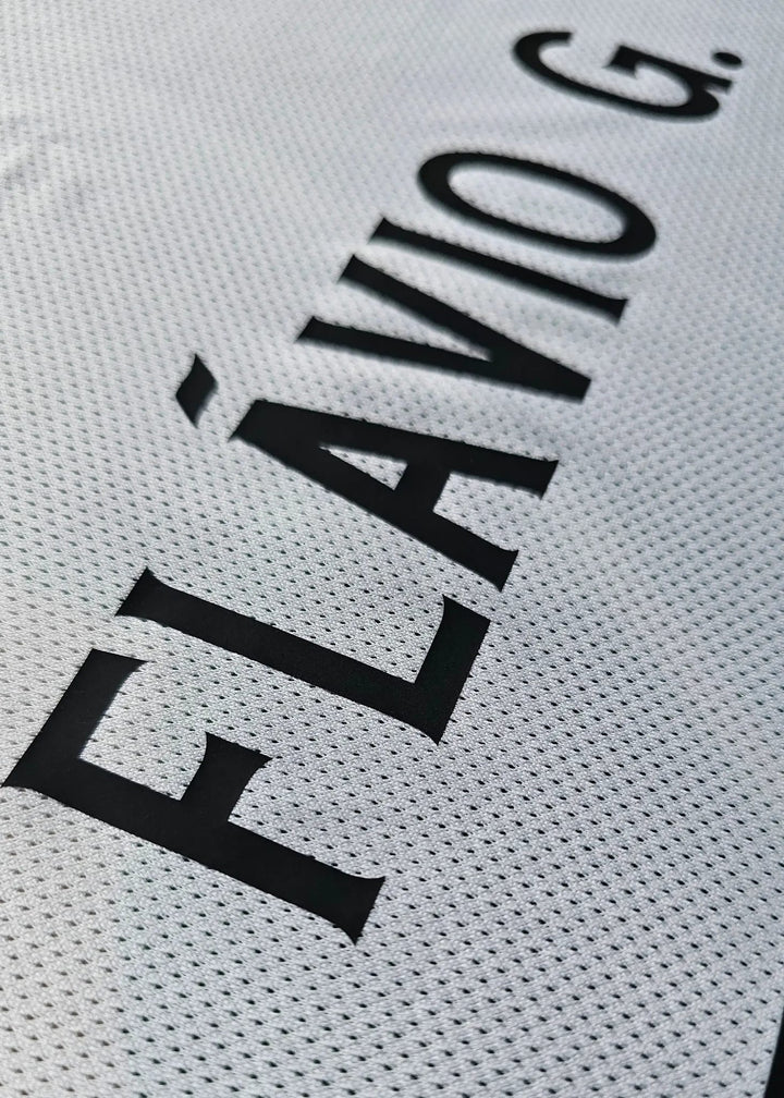 Close-up of a white perforated athletic jersey with bold black text reading 'FLÁVIO G.' printed diagonally across the fabric.