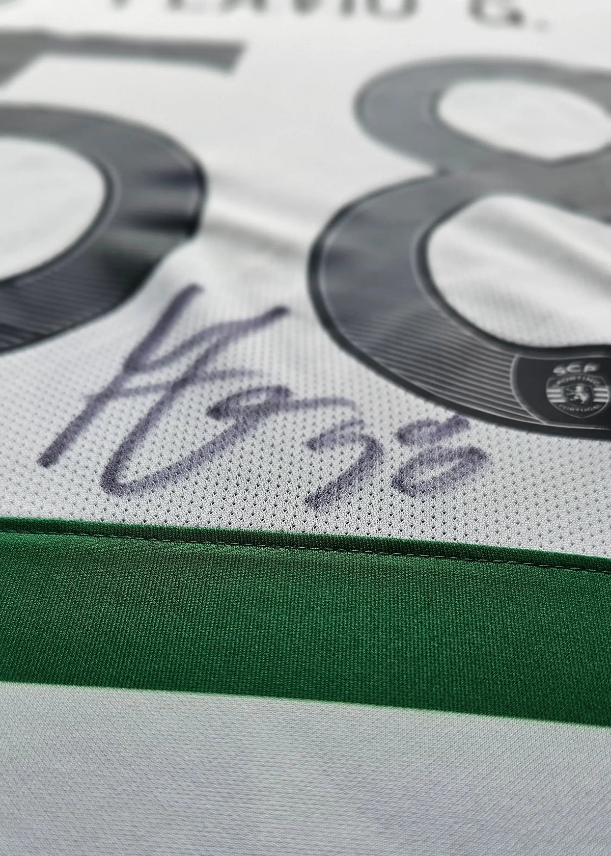 Close-up of a white soccer jersey with a green horizontal band, large dark gray number 58, and a dark signature on the front.