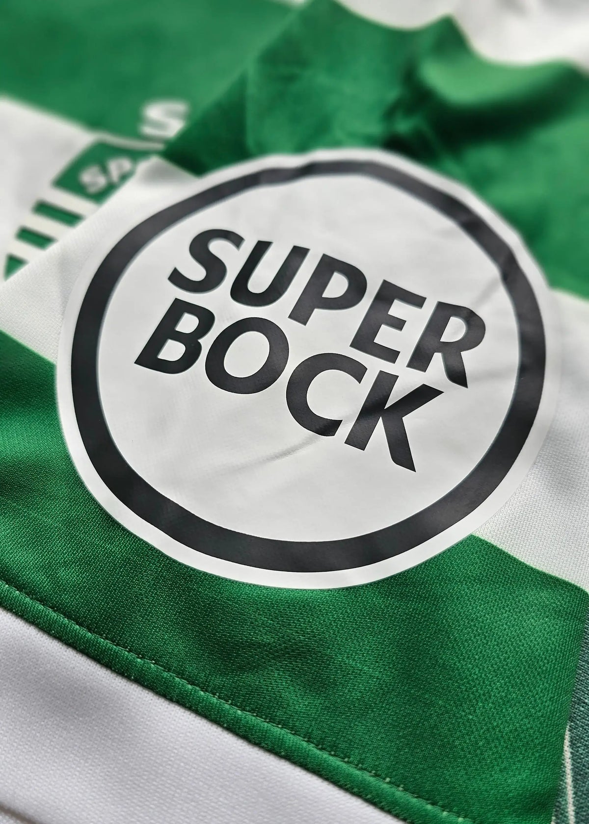 Close-up of the round black and white Super Bock sponsor patch on green and white striped fabric, Flavio Gonçalves' Sporting CP shirt.
