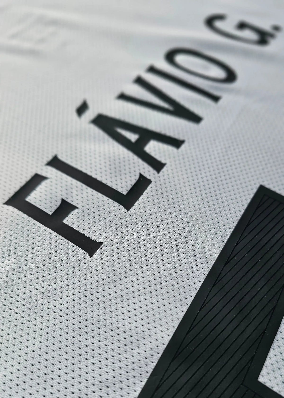 Close-up of a white soccer jersey with black lettering reading 'FLÁVIO G.' and a large black number block, showing the textured mesh fabric.