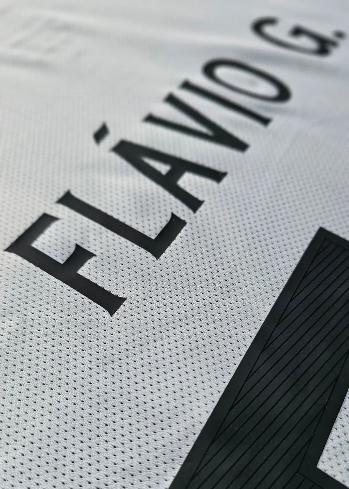 Close-up of a white soccer jersey with black lettering reading 'FLÁVIO G.' and a large black number block, showing the textured mesh fabric.