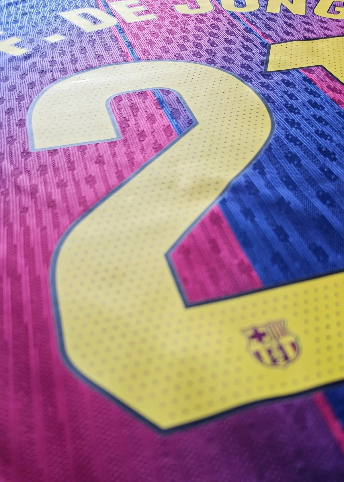 Yellow 'DE JONG' nameset and a corner of the gold FC Barcelona crest, above the number 21, on Frenkie de Jong's purple-pink gradient shirt back.